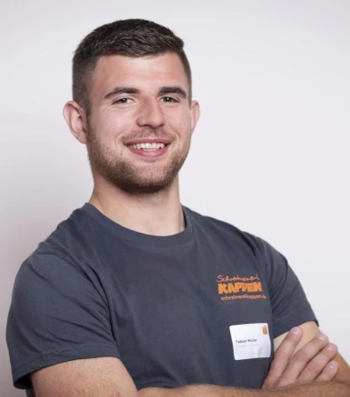 Junger Handwerker mit T-Shirt und Namensschild lächelt vor neutralem Hintergrund.