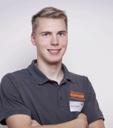 Junger Mann im grauen Firmen-Poloshirt mit verschränkten Armen vor hellem Hintergrund.