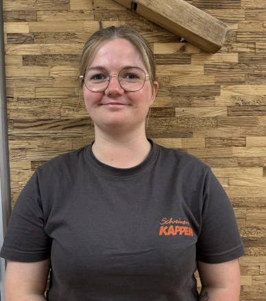 Junge Frau mit Brille und T-Shirt „Schreinerei Kappen“ vor Holzdekor-Wand.