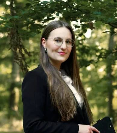 Junge Frau mit Brille und langen Haaren im Business-Outfit im grünen Wald stehend.