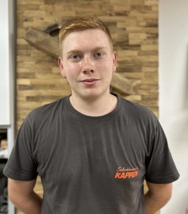 Junger Handwerker mit braunem T-Shirt steht vor Holzwand in moderner Schreinerei.