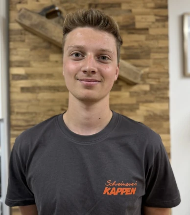 Junger Mann im T-Shirt der Schreinerei Kappen steht vor holzverkleideter Wand.