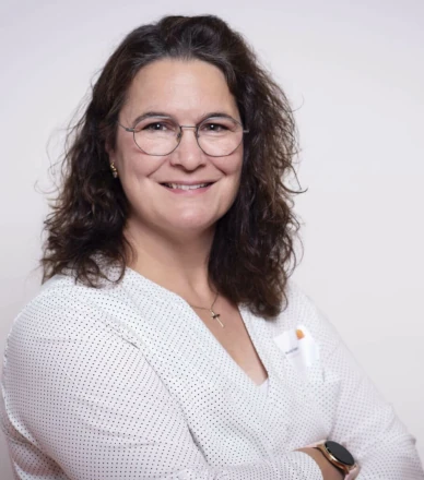 Frau mit lockigen braunen Haaren und Brille lächelt im weißen Hemd vor hellem Hintergrund.