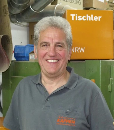 Älterer Mann in Werkstatt mit grauem Haar, lächelnd vor Schrank und Tischler-Karton.