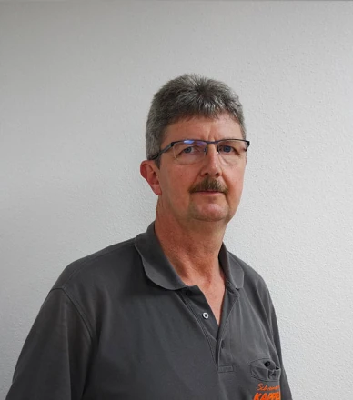 Mann mit Brille und grauem Haar steht vor weißer Wand und blickt in die Kamera.